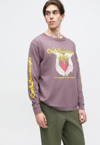 Ilgas rankovės violetinis marškinis, pagamintas iš minkštos medžiagos, su rankų, laikančių širdį su liepsnomis, grafiniu dizainu ir geltonu tekstu ant rankovių.