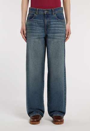 Person wearing loose-fitting blue jeans and brown shoes, standing against a plain light background.