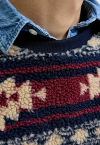 Fleece trui met een gestructureerd oppervlak met rode, blauwe en crèmekleurige geometrische patronen. Afgewerkt met een marineblauw ribbelkraag onder een denim shirt.