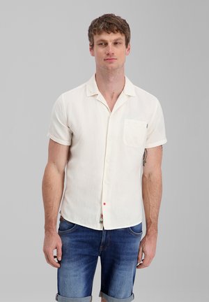 Mann trägt kurzärmliges weißes Hemd und blaue hochgekrempelte Jeansshorts, steht vor einfarbigem grauem Hintergrund.