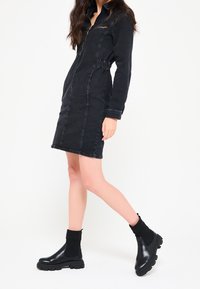 Musta farkkukleid, jossa on pitkät hihat, vetoketjullinen etuosa ja kapea vyötärö. Yhdistetty paksujen mustien nilkkabootsien ja ribbisaappaiden kanssa.