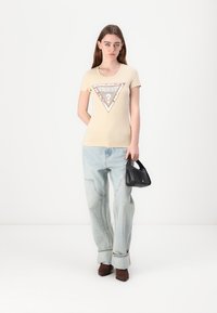 T-shirt ajustada bege com um gráfico triangular em prata e branco, combinada com jeans baggy azul claro e uma bolsa preta. Sapatos de salto marrons.