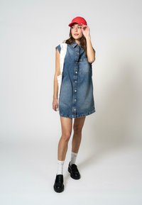 Farmerdzseki ruházat gombos elejével, rövid ujjakkal és mellzsebbe zsebével. Piros sapkával, fekete loaferekkel és fehér zoknikkal viselve.