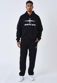 Schwarzes Zip-Hoodie und Sweatpants-Set, das ein großes weißes Logo auf der Brust und ein kleineres Logo am Hosenbein aufweist. Kombiniert mit weißen Sneakern.