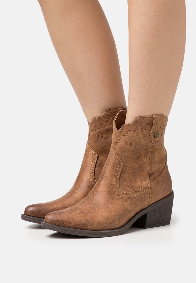 Bottes en daim marron mi-mollet avec un talon bas empilé et des coutures décoratives portées sur jambes nues.