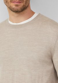 Beżowy sweter dzianinowy z okrągłym dekoltem i długimi rękawami, o delikatnej fakturze i ściągaczach przy rękawach. Nałożony na biały T-shirt.