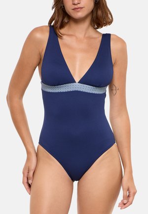 Femme portant un maillot de bain une pièce texturé bleu marine avec un décolleté en V profond et une bande à motifs sous la poitrine sur fond blanc.