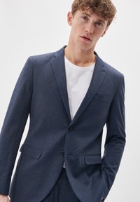 Navy blue blazer in textured fabric with two button closure, notch lapels, and front pockets. Worn over a white t-shirt.