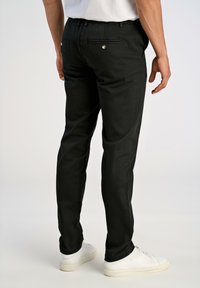 Hombre con pantalones slim-fit negros y zapatillas blancas, visto desde atrás sobre un fondo liso.