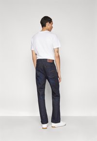 Mørkeblå denim jeans med lige pasform, brun læderpatch på ryggen og kontrastsyede stikninger. Bæres med en simpel hvid t-shirt.