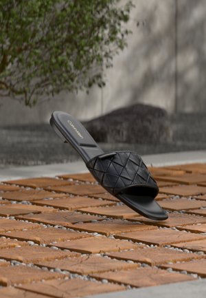 Fekete Calvin Klein papucs, steppelt pánttal, fa járólapon kültéren, zöld növényekkel és kőfalas háttérrel.