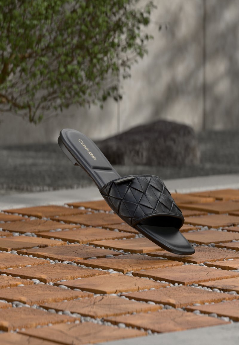 Μαύρο σανδάλι τύπου slide Calvin Klein με επάνω μέρος από καπιτονέ ύφασμα, τοποθετημένο πάνω σε ξύλινα πλακάκια σε εξωτερικό χώρο με φόντο πράσινο φυλλώδες τοπίο και πέτρινο τοίχο.