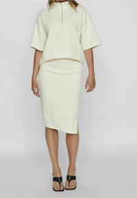 Ensemble léger crème en deux pièces comprenant un haut à manches courtes avec un col zippé et une jupe midi avec une fente sur le côté, complété par des sandales noires.