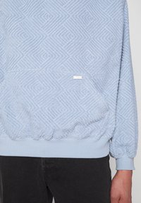 Sweat-shirt bleu clair texturé avec des motifs géométriques et une poche avant, porté avec un pantalon noir, montrant le bas du torse et la main droite.