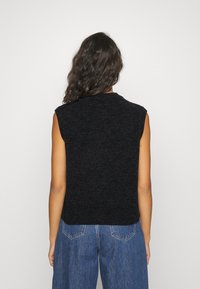 Donna con lunghi capelli ricci, che indossa un top in maglina nera senza maniche e jeans blu larghi, in piedi di fronte a uno sfondo bianco semplice.