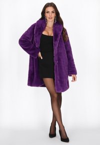 Een vrouw in een paars nep bontjack over een zwart mini-jurk, met sheer zwarte panty’s, zwarte hakken en lange oorbellen, staat met één hand in haar zak.