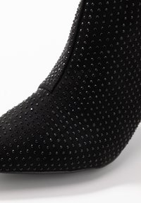 Zapato de tacón alto de ante negro con punta afilada, cubierto de pequeños rhinestones negros brillantes, fotografiado sobre un fondo blanco.