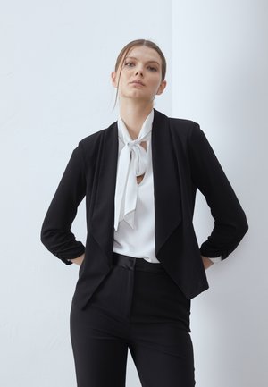 Blazer nero su misura con maniche lunghe, design aperto davanti, abbinato a una camicetta bianca con sciarpa annodata al collo. Tessuto morbido, stile minimalista.