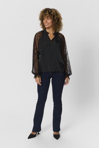 Juoda bluzonė su nėrinių rankovėmis, V formos iškirpte ir šoniniu raukiniu, derinama su tamsiai mėlynomis džinsomis ir juodais, smailiais bateliais.