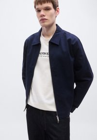 Μπλε ναυτικό μπουφάν με φερμουάρ και επίπεδη γιακά, κατασκευασμένο από λείο ύφασμα, φορεμένο πάνω από λευκό φούτερ με στρογγυλή λαιμόκοψη, σε συνδυασμό με σκούρο τζιν.