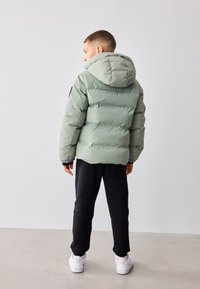 Jachetă pufoasă de culoare verde deschis, cu glugă, având secțiuni quilted și un accent negru pe mânecă. Asortată cu pantaloni sport negri și adidași albi.