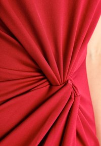 Röd tyg med samlad textur, med en centralt vriden detalj. Slätt stickat material, närbild som betonar veck och draperingar.