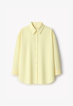 Chemise à manches longues jaune pâle avec col classique et poignets boutonnés, présentée sur un fond blanc.