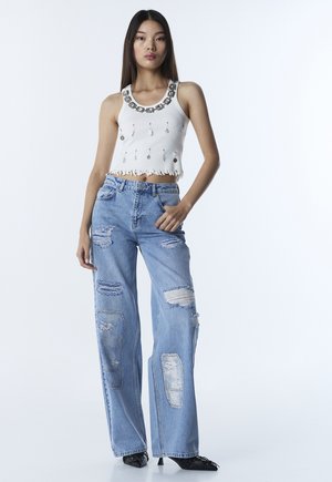 Mujer joven con top corto sin mangas blanco adornado y jeans anchos de pierna ancha y desgastados en azul claro, con tacones negros puntiagudos.