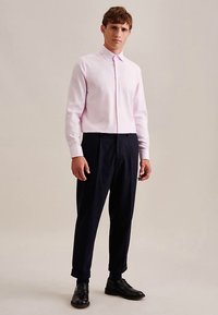 Camicia a maniche lunghe rosa chiaro con colletto con bottoni, abbinata a pantaloni blu navy con taglio affusolato e scarpe nere, su uno sfondo neutro.