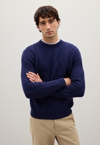 Marineblauer gestrickter Pullover mit Rautenmuster, geripptem Rundhalsausschnitt und Bündchen. Das Modell trägt beigefarbene Hosen, hat die Arme verschränkt und steht vor einem neutralen Hintergrund.