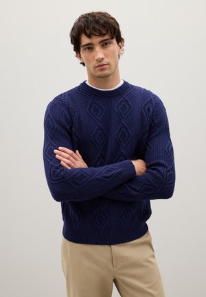 Marineblauer gestrickter Pullover mit Rautenmuster, geripptem Rundhalsausschnitt und Bündchen. Das Modell trägt beigefarbene Hosen, hat die Arme verschränkt und steht vor einem neutralen Hintergrund.