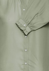 Light green blouse with a smooth texture, featuring button closures and ruffled sleeve cuffs, highlighting a classic design.