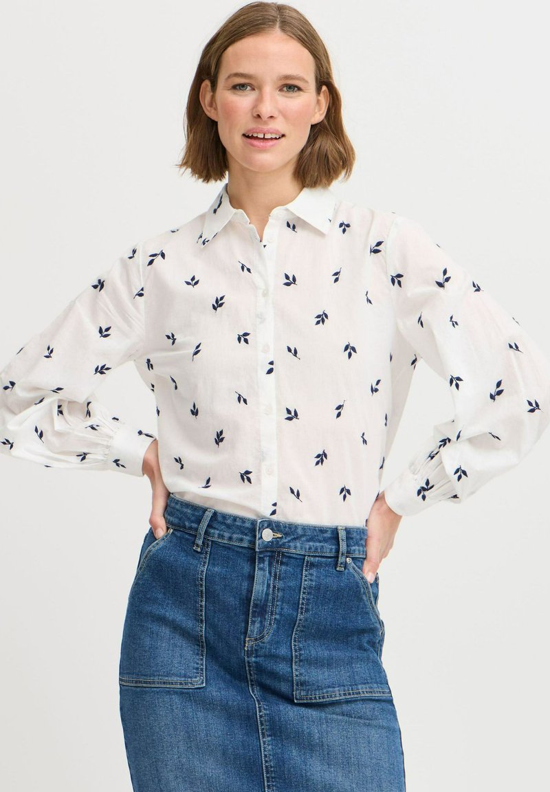 Weißes Hemd mit navyblauem Blättermuster, knöpfbarem Design und langen Ärmeln. Kombiniert mit einem blauen Jeansrock mit Fronttaschen.
