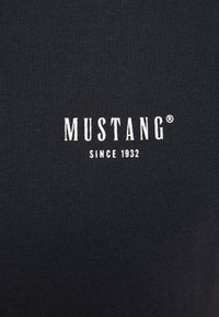 Fekete pamut póló bordázott textúrával és középen elhelyezett fehér nyomott logóval, amely így olvasható: "MUSTANG SINCE 1932".