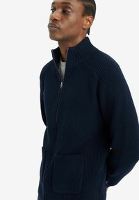 Suéter azul marino de cremallera con textura acanalada, cuello alto y dos bolsillos frontales; presenta un corte relajado y material de punto grueso.