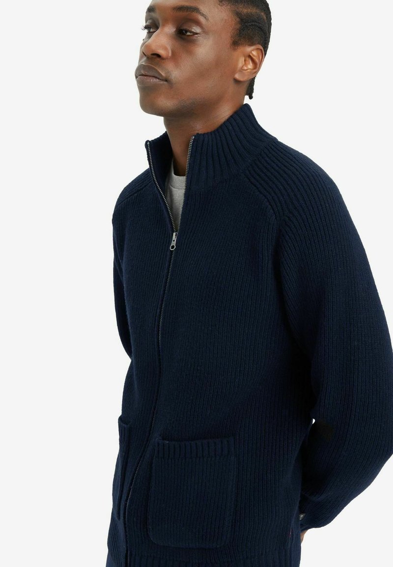 Suéter azul marino de cremallera con textura acanalada, cuello alto y dos bolsillos frontales; presenta un corte relajado y material de punto grueso.