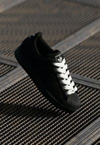 Svart sneaker med hvite snørebånd, har en teksturert overdel, tre striper og en gummiskalltå. Merkevaren sitt logo er synlig på tungen.