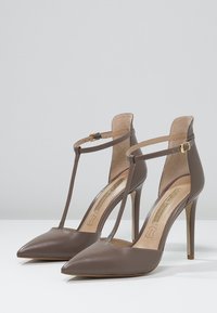 Chaussures à talons hauts en cuir verni marron avec bouts pointus, design à sangle en T, et accent en boucle dorée. Doublure intérieure texturée.