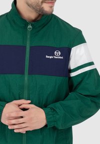 Grüne Nylonjacke mit einem marineblauen Bruststreifen, weißen horizontalen Akzenten und elastischen Bündchen. Verfügt über das Sergio Tacchini-Logo auf der Brust.