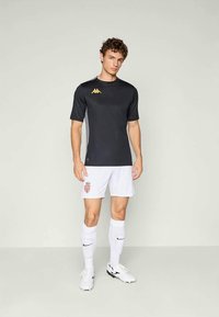 Must-katte lühikeste varrukatega sportlik särk, millel on kuldne logo, koos valgete lühikesed pükstega, millel on punane embleem. Mudel kannab musta ja valget sportlikku jalanõud.