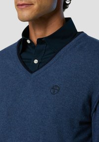 Blauer V-Ausschnitt-Pullover aus strukturiertem Strickstoff, getragen über einem dunklen Hemd mit Kragen. Mit einem dezenten Logo auf der linken Brustseite.
