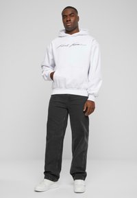 Sudadera blanca con bolsillo frontal y logo de script negro, combinada con pantalones sueltos gris oscuro y zapatillas blancas. Tela suave, diseño casual.
