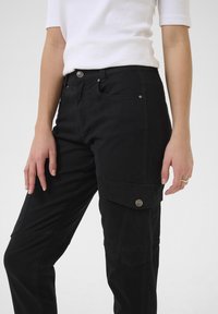 Pantalons cargo noirs avec poches à fermetures à pression. Fabriqués en tissu texturé, conçus avec une coupe ajustée et une taille mi-haute.