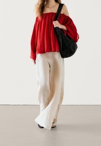 Rød off-shoulder bluse med løstsiddende snit og plisseringer, parret med cremefarvede vidde bukser. Modellen holder en sort ruskinds taske med skulderrem.