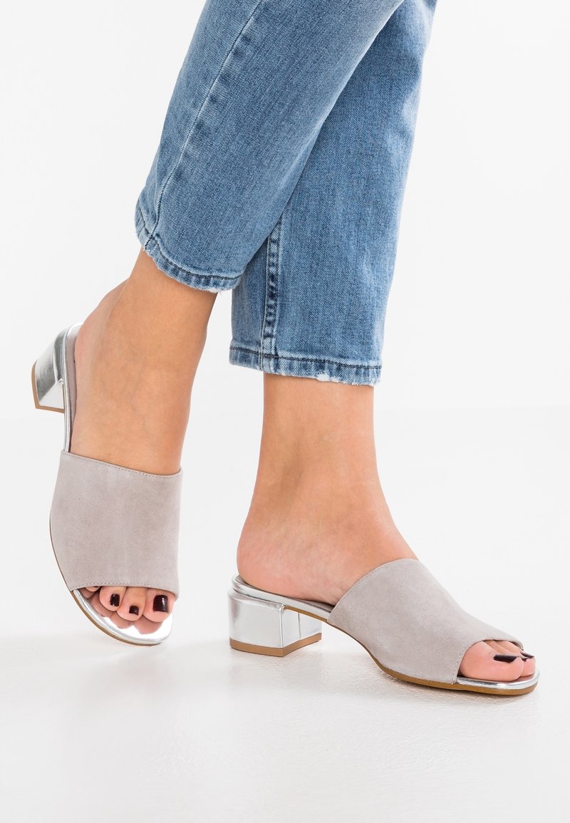 Mules à bout ouvert en daim gris avec un petit talon carré argenté. Texture lisse et design minimaliste ; esthétique chic et moderne.