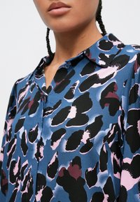 Skjorte med knapper fremme, med et abstrakt dyreprint i blått, svart og rosa. Den har spredt krage og en teksturert stoffoverflate.