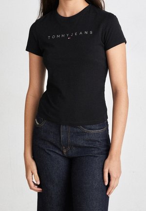 Kobieta w dopasowanym, czarnym t-shircie Tommy Jeans z krótkim rękawem i ciemnoniebieskich dżinsach na jednolitym tle.