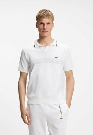 Giovane uomo con capelli biondi corti che indossa una polo bianca BOSS con dettaglio in mesh e pantaloni bianchi abbinati con striscia nera e dorata.