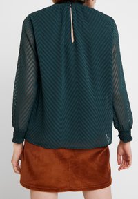 Blouse en chiffon vert avec un motif en chevrons, encolure froncée et poignets élastiques ; détail d'ouverture en forme de clé au dos visible. Associe avec une jupe en velours côtelé marron.