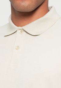 Gros plan d'une personne portant un polo couleur crème avec une patte à deux boutons et un col doux et arrondi.